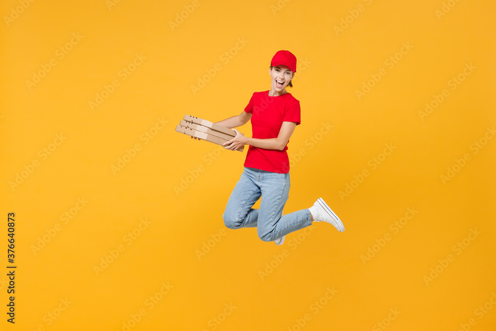 Full length body fun delivery employee woman in red cap blank t-shirt uniform work courier in service jumping hold food order pizza in cardboard flatbox isolated on yellow background studio portrait. Full length body fun delivery employee woman in red cap blank t-shirt uniform work courier in service jumping hold food order pizza in cardboard flatbox isolated on yellow background studio portrait.