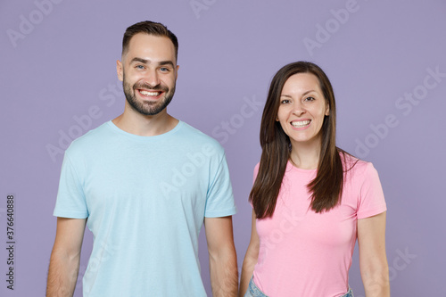 Smiling cheerful funny beautiful young couple two friends man woman 20s wearing blue pink empty blank design t-shirts stand looking camera isolated on pastel violet color background studio portrait.