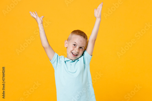 Smiling happy little fun male kid boy 5-6 years old in stylish blue turquoise t-shirt polo raised hands up, celebrating doing winner gesture isolated on yellow color background, child studio portrait.