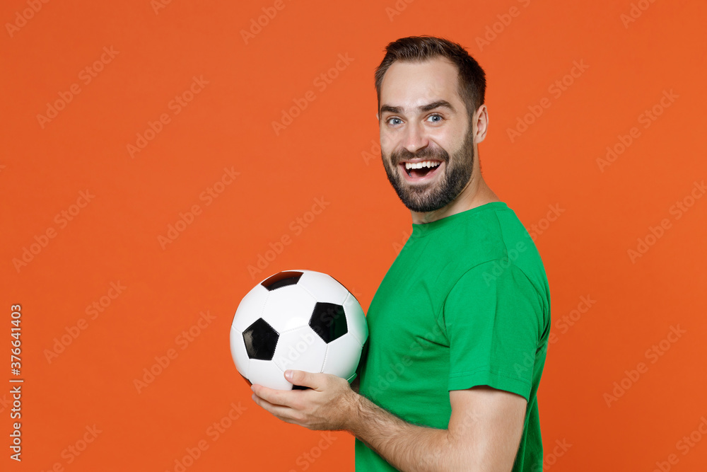 Side view excited young man football fan in green t-shirt cheer up support favorite team with soccer ball looking camera isolated on orange background studio. People sport leisure lifestyle concept.