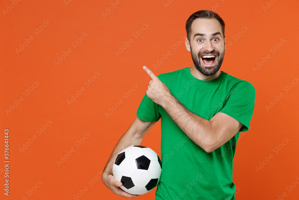 Excited man football fan in green t-shirt cheer up support favorite team with soccer ball point index finger aside up on mock up copy space isolated on orange background. People sport leisure concept. Excited man football fan in green t-shirt cheer up support favorite team with soccer ball point index finger aside up on mock up copy space isolated on orange background. People sport leisure concept.