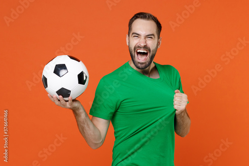 Cheerful young man football fan tearing green t-shirt on himself cheer up support favorite team with soccer ball isolated on orange background studio portrait. People sport leisure lifestyle concept.