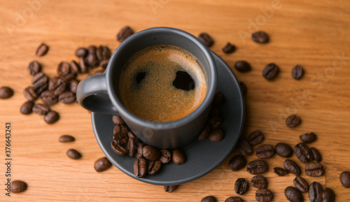 A grey cup of coffee on a wooden table with fresh coffee beans around with against a dark background