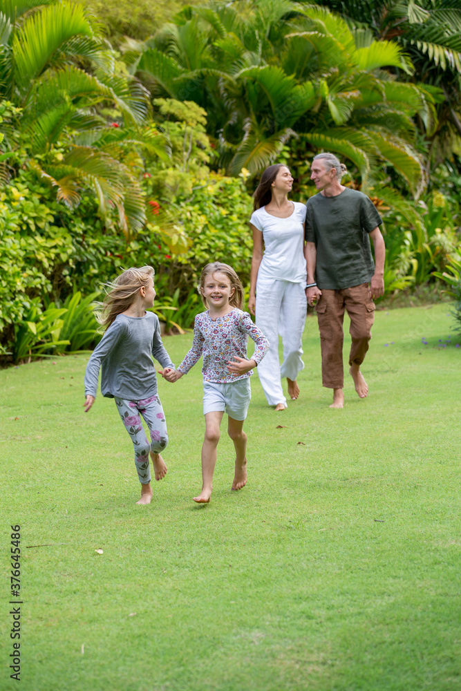 Fototapeta premium Young happy family walking in tropical park or gaden with palm trees. Two cute little sisters running in front of parents. Summer exotic vacation