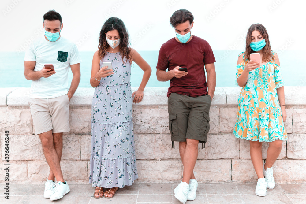 Friends group using smartphone leaning against the wall - Young people with surgical mask keeps social distancing. Friends group using smartphone leaning against the wall - Young people with surgical mask keeps social distancing.
