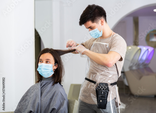Positive male hairdresser in protective mask and gloves making hair styling for young woman  working day in beauty salon during pandemic situation