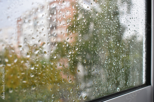 Raindrops on a rainy day on the window pane. Autumn cloudy weather through glass covered with rain drops. Blurred autumn cityscape. Selective focus on raindrops.