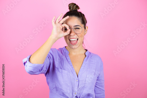 Young beautiful woman wearing glasses over isolated pink background doing ok gesture shocked with smiling face  eye looking through fingers