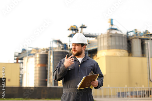 American engineer holding walkie talkie and notes, factory in background.