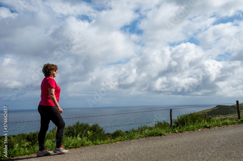 Wide shot of mature woman walking along a country road with the sea in the background and cloudy sky, horizontal, with copy space © Arantxa Forcada