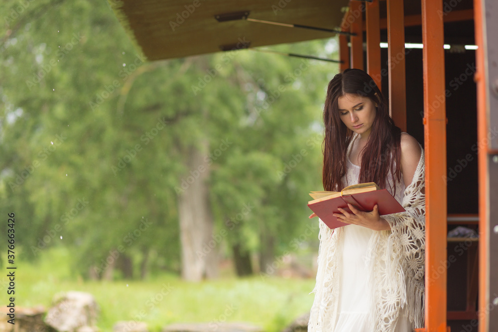Naklejka premium Pretty woman in white dress reading book in the cloudy nature. Girl sits on the terrace or on the porch