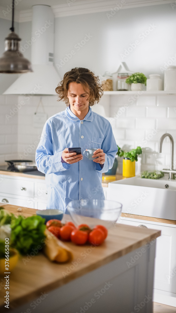 Happy Young Man with Long Hair Using Smartphone while Wearing Blue Pajamas. He is Scrolling News Feed, Social Media, Chatting and Drining Morning Coffe in a Kitchen with Healthy Vegetables. Happy Young Man with Long Hair Using Smartphone while Wearing Blue Pajamas. He is Scrolling News Feed, Social Media, Chatting and Drining Morning Coffe in a Kitchen with Healthy Vegetables.