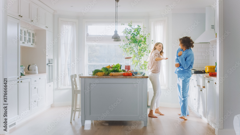 Handsome Young Man in Blue Pajamas and Beautiful Girl in Striped Jumper are Fooling Around in the Kitchen. White Modern Kitchen Area with Healthy Green Vegetables on a Table. Happy Couple in Cozy Home Handsome Young Man in Blue Pajamas and Beautiful Girl in Striped Jumper are Fooling Around in the Kitchen. White Modern Kitchen Area with Healthy Green Vegetables on a Table. Happy Couple in Cozy Home