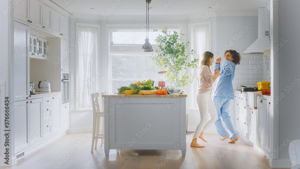 Handsome Young Man in Blue Pajamas and Beautiful Girl in Striped Jumper are Fooling Around in the Kitchen. White Modern Kitchen Area with Healthy Green Vegetables on a Table. Happy Couple in Cozy Home Handsome Young Man in Blue Pajamas and Beautiful Girl in Striped Jumper are Fooling Around in the Kitchen. White Modern Kitchen Area with Healthy Green Vegetables on a Table. Happy Couple in Cozy Home