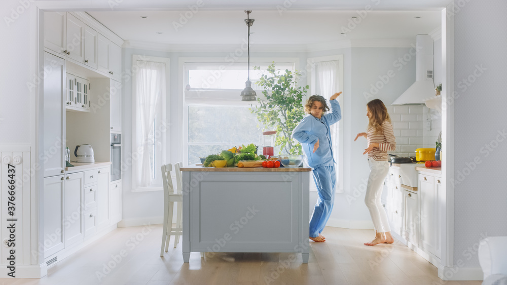 Handsome Young Man in Blue Pajamas and Beautiful Girl in Striped Jumper are Dancing in the Kitchen. Bright White Modern Kitchen Area with Healthy Green Vegetables on a Table. Happy Couple in Cozy Home Handsome Young Man in Blue Pajamas and Beautiful Girl in Striped Jumper are Dancing in the Kitchen. Bright White Modern Kitchen Area with Healthy Green Vegetables on a Table. Happy Couple in Cozy Home