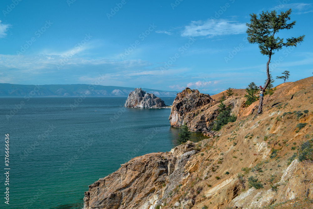 Cape Shamanka rock in the clear blue Lake Baikal among the grassy steppes with coniferous larch trees, against the background of mountains and clouds, blue sky