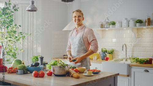 Handsome Man in Pink Shirt and Apron is Making a Healthy Organic Salad Meal in a Modern Sunny Kitchen. Hipster Man in Glasses Smiles at the Camera. Natural Clean Diet and Healthy Way of Life Concept.