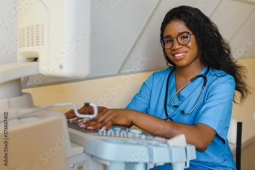 Young female African-american doctor working on modern ultrasound equipment. Operator of ultrasound scanning machine sitting and looking at the monitor, waiting for patient.