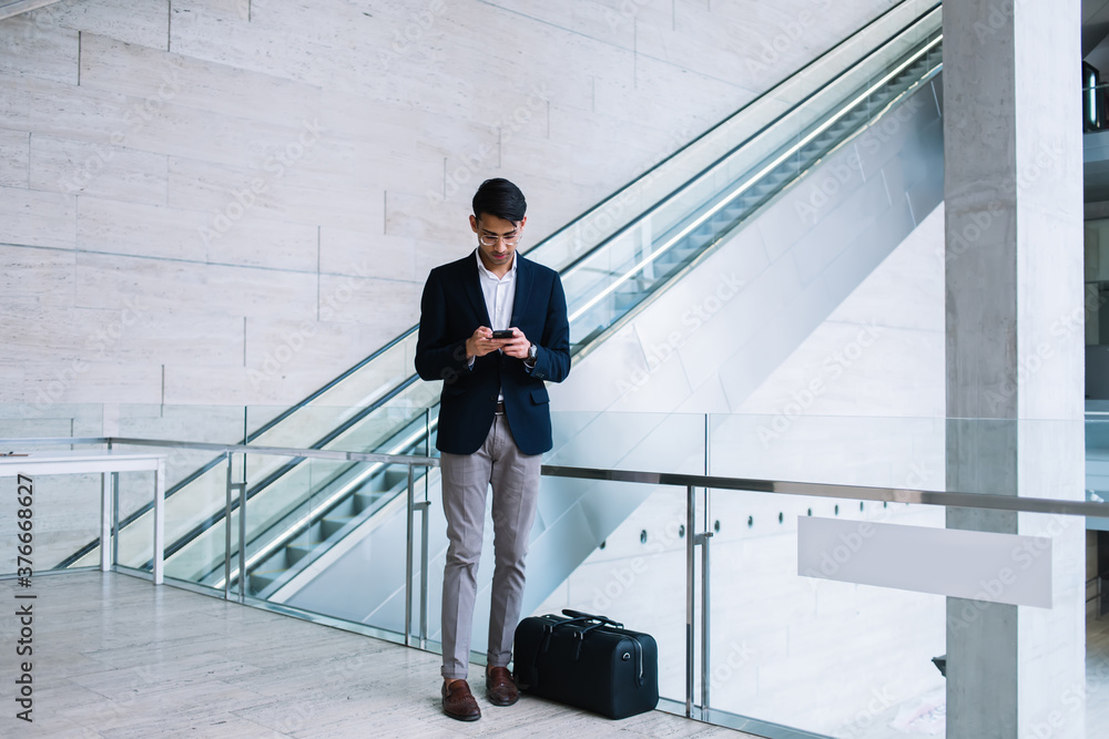 Handsome prosperous hindu businessman using mobile phone for dialling number standing near bag for trip, serious prosperous male intelligent proud ceo reading news on smartphone near luggage Handsome prosperous hindu businessman using mobile phone for dialling number standing near bag for trip, serious prosperous male intelligent proud ceo reading news on smartphone near luggage