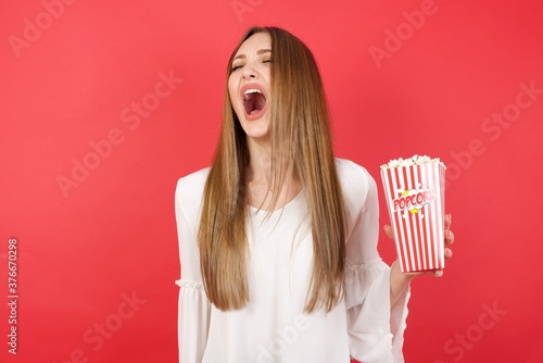 Eastern European woman holding bucket with popcorn standing over isolated red background angry and mad screaming frustrated and furious, shouting with anger. Rage and aggressive concept.