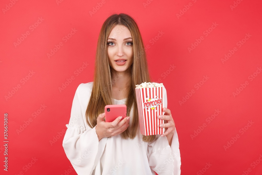 Eastern European woman holding bucket with popcorn standing enjoys distant communication, uses mobile phone, surfs fast unlimited internet, has pleasant smile, makes shopping online, Eastern European woman holding bucket with popcorn standing enjoys distant communication, uses mobile phone, surfs fast unlimited internet, has pleasant smile, makes shopping online,
