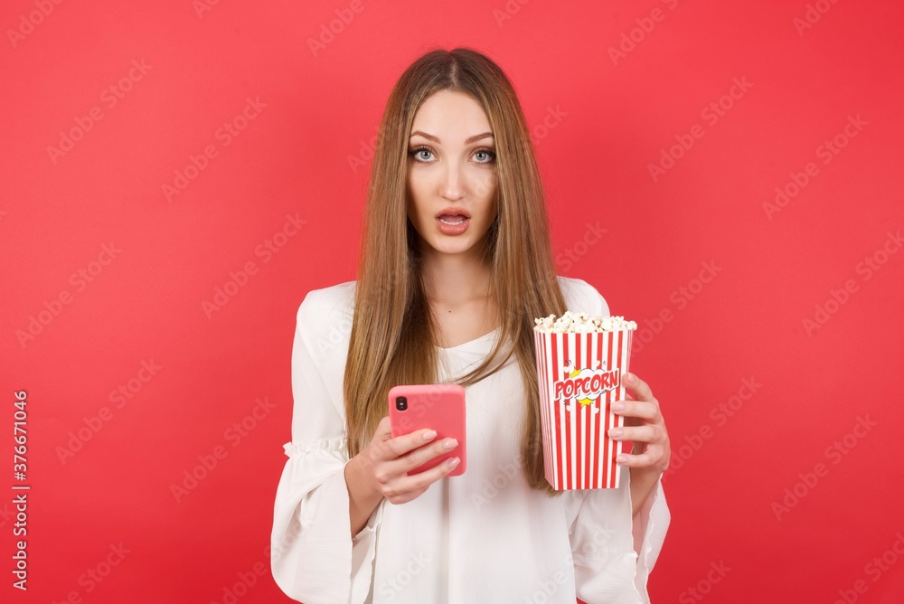 Eastern European woman holding bucket with popcorn standing over isolated red background. opens mouth hold telephone reading advert unbelievable big shopping prices Eastern European woman holding bucket with popcorn standing over isolated red background. opens mouth hold telephone reading advert unbelievable big shopping prices
