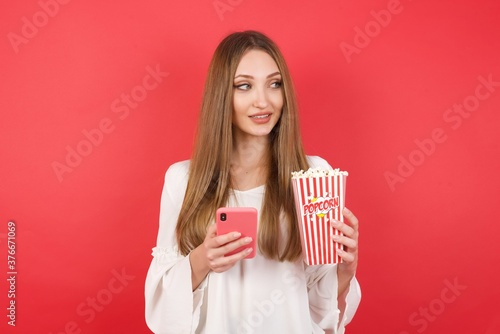 Eastern European woman holding bucket with popcorn standing over isolated red background hold telephone hands read good youth news look empty space advert
