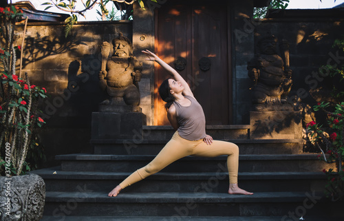 Positive caucasian female in tracksuit stretching muscles standing in asana on practice during vacation retreat, calm concentrated woman enjoying relaxation and vitality on yoga training near temple