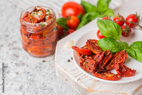 Sundried tomatoes on the white plate and in a glass jar on a white wooden board on a gray background. Next to the fresh tomatoes and green basal. Close-up, space for text, copy space