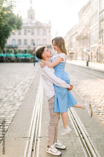 Adorable couple in love, enjoying their walk on a sunny day in the city, standing on tram track. Handsome guy holds his attractive young girldriend in blue dress on hands