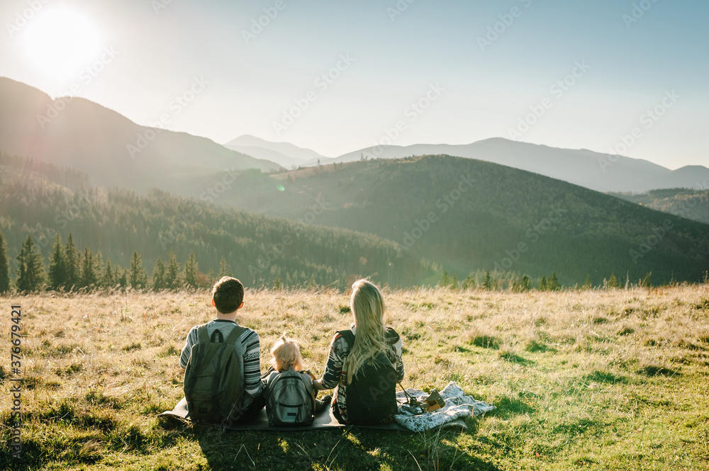 Mom, dad and daughter in the mountains enjoy sunset and look at nature. Back view. Young family spending time together on vacation, outdoors. The concept of family autumn holiday.