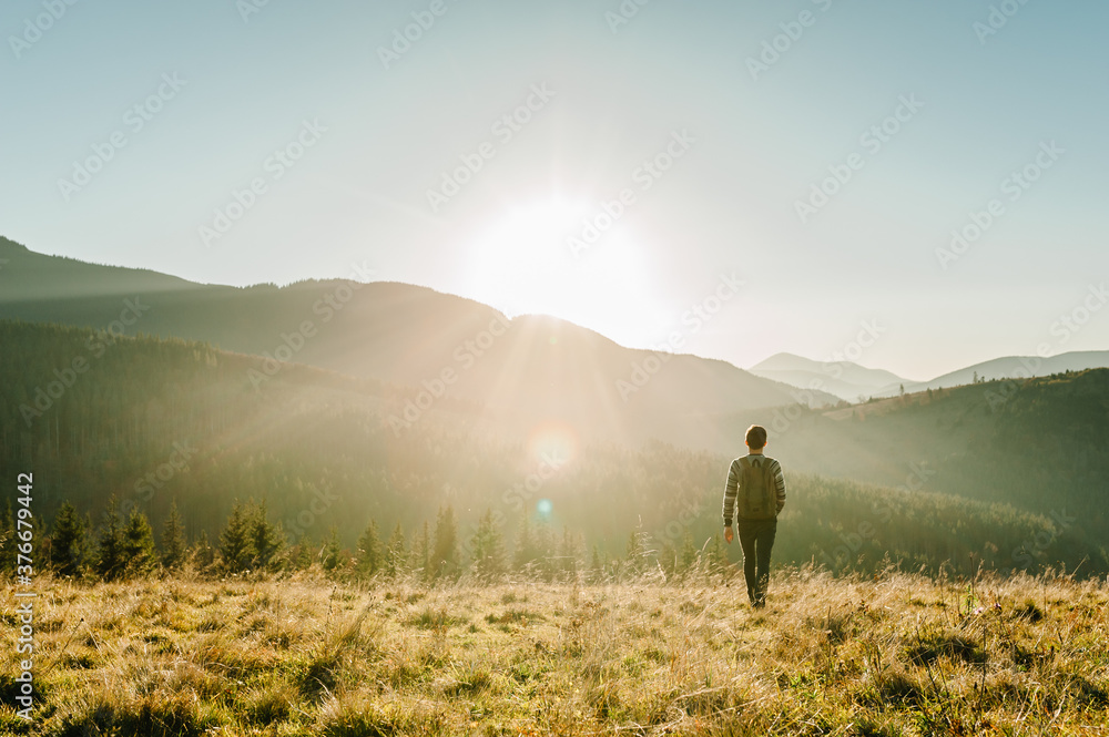 A young tourist man with a backpack on top and enjoying the sunset. Back view. Hipster walks in the autumn grass in the mountains on vacation. Holiday trip concept. World Tourism Day.