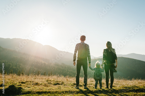 Family with kid hiking in mountains. Young tourists on top of a mountain enjoying valley view sunset. Happy mom, dad with a backpack and daughter. Holiday trip concept. World Tourism Day. Back view.