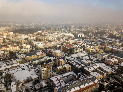 Aerial black and white winter top view of modern city center with tall buildings and parked cars on snowy streets.