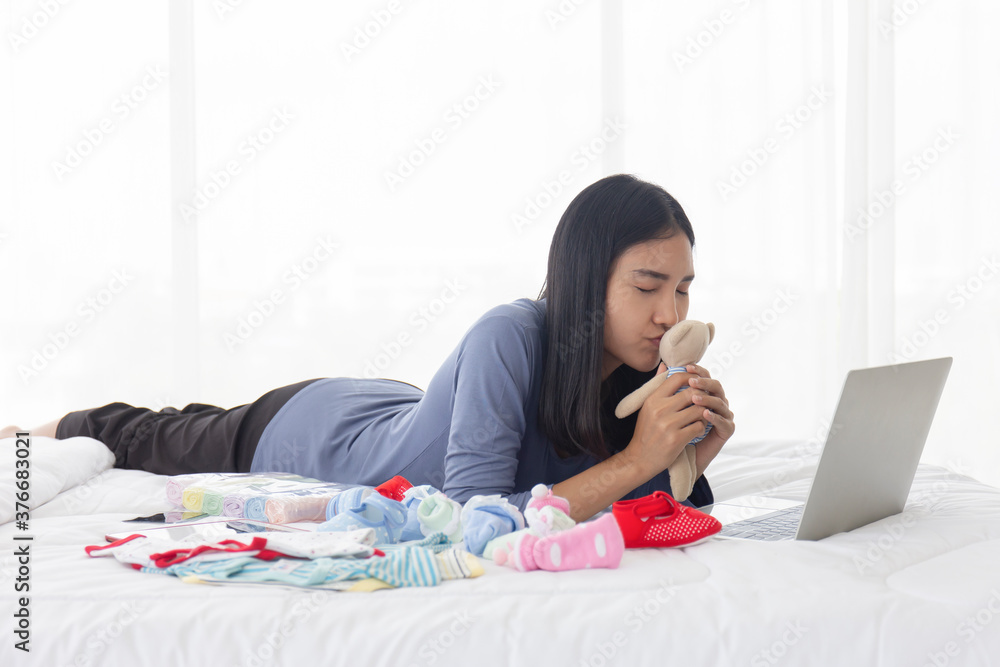 An Asian mom kissing her baby's unused teddy bear to sell online in a laptop - second-hand good in bed. An Asian mom kissing her baby's unused teddy bear to sell online in a laptop - second-hand good in bed.