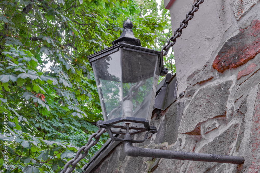 Street old lantern, made of metal, suspended on a stone wall, in the territory of a medieval fortress, one cloudy, summer day