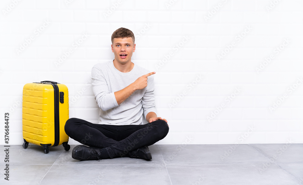 Young handsome man sitting on the floor with a suitcase pointing finger to the side Young handsome man sitting on the floor with a suitcase pointing finger to the side