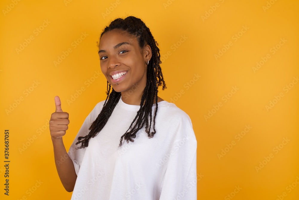 Young dark skinned woman with braids hair wearing white t-shirt over yellow background doing happy thumbs up gesture with hand. Approving expression looking at the camera showing success. Young dark skinned woman with braids hair wearing white t-shirt over yellow background doing happy thumbs up gesture with hand. Approving expression looking at the camera showing success.