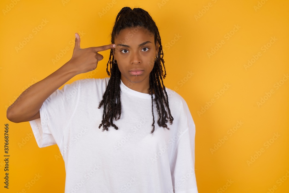 Unhappy Young dark skinned woman with braids hair wearing white t-shirt over yellow background. curves lips and makes suicide gesture, tired of everything, shoots in temple with hand, Unhappy Young dark skinned woman with braids hair wearing white t-shirt over yellow background. curves lips and makes suicide gesture, tired of everything, shoots in temple with hand,