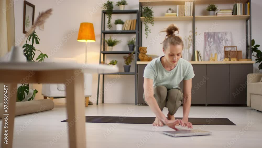Tracking shot of young woman in sportswear opening laptop and sitting down on fitness mat to practice yoga with online instructor during pandemic Tracking shot of young woman in sportswear opening laptop and sitting down on fitness mat to practice yoga with online instructor during pandemic