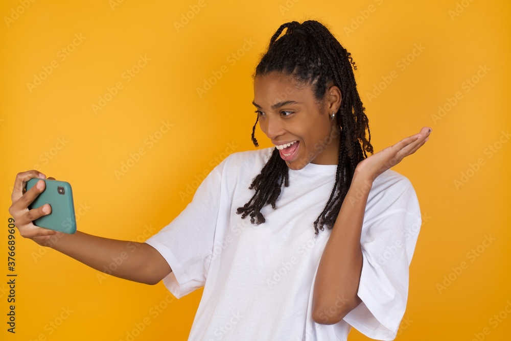 Wow!! excited Young dark skinned woman with braids hair wearing casual clothes trendy isolated over yellow background showing mobile phone with open hand gesture Wow!! excited Young dark skinned woman with braids hair wearing casual clothes trendy isolated over yellow background showing mobile phone with open hand gesture