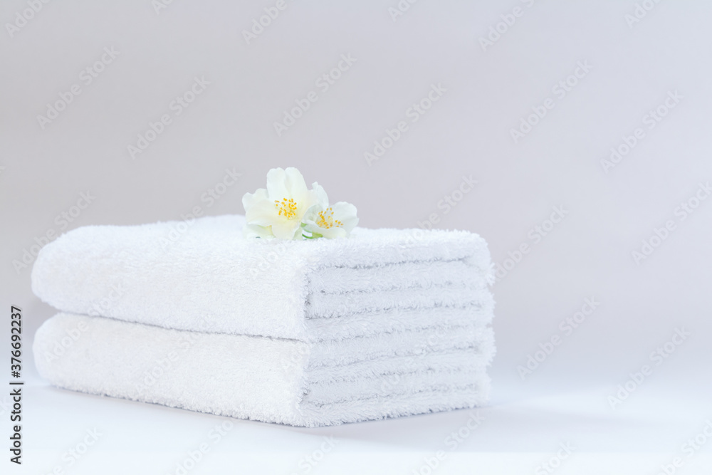 Two white neatly folded terry towels with a jasmine flower on a light background. Two white neatly folded terry towels with a jasmine flower on a light background.