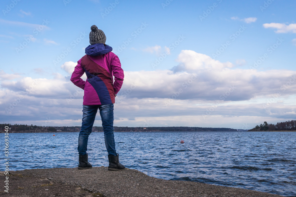 Young woman on rocky coast looking at the sea. Autumn Fall Concept. View of Scandinavian Stockholm Archipelago on cloudy sky background. Pine forest islands on the horizon.