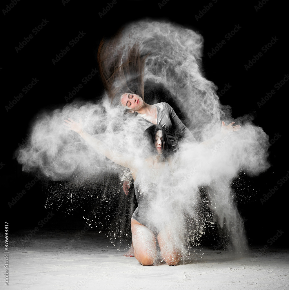 two beautiful young caucasian women in black bodysuits with a sports figure are dancing in a white cloud of flour