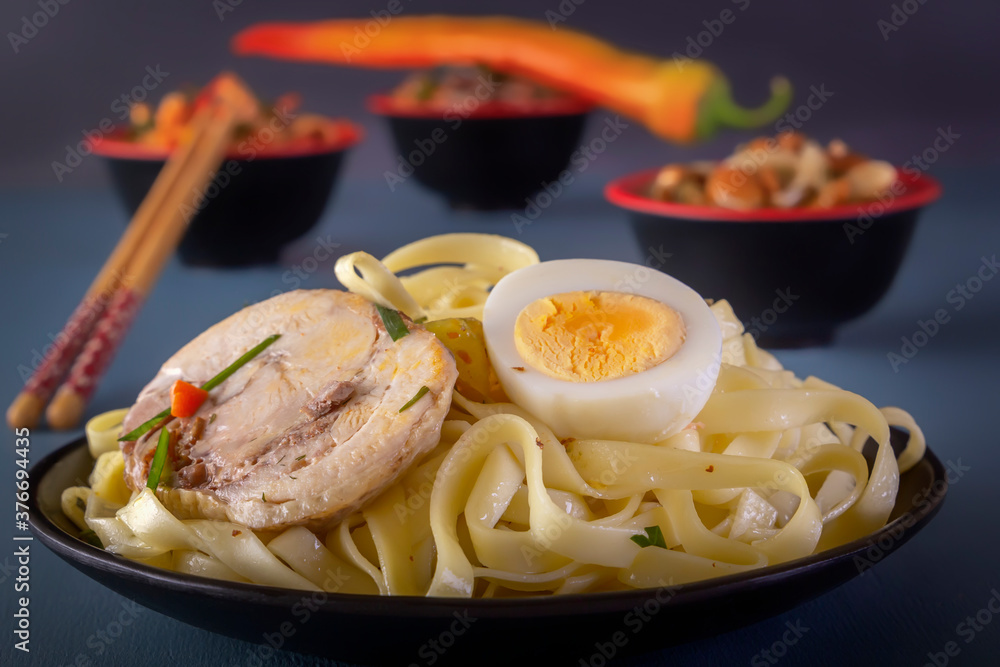Noodles with pieces of meat and eggs in a black plate and dim sum with various traditional mushroom, soy and vegetable snacks with chopsticks on a blue table. Asian food concept Noodles with pieces of meat and eggs in a black plate and dim sum with various traditional mushroom, soy and vegetable snacks with chopsticks on a blue table. Asian food concept