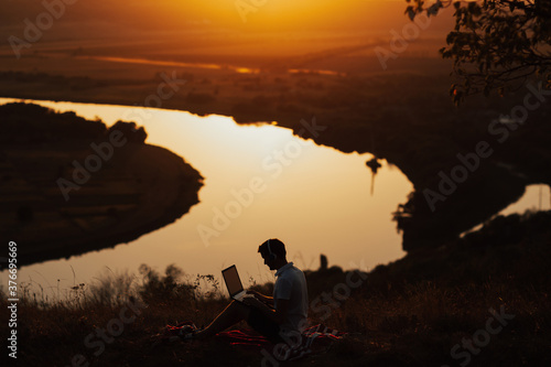 Silhouette of man working on laptop, he sitting on the peak of a mountain at sunset with river on background. Technology, internet, staying online in nature.
