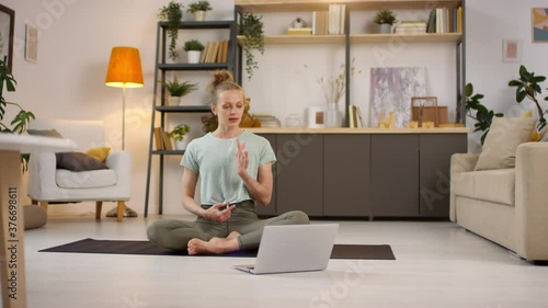 Tracking of young female yoga instructor sitting on mat at home and looking at laptop while talking and explaining poses during online class photo