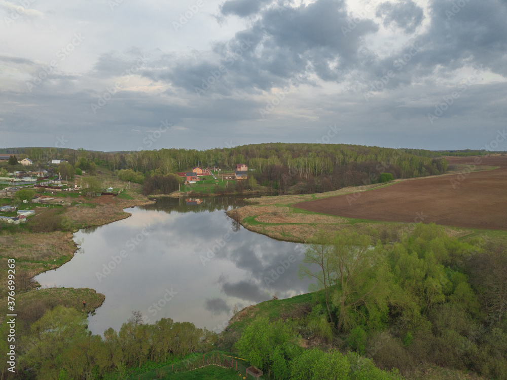 Obraz premium Aerial perspective view of the pond surrounded by settlements, fields and forests under cloudy sky. Drone air view of the agricultural landscape with lake, village, trees and meadows. Summer day.