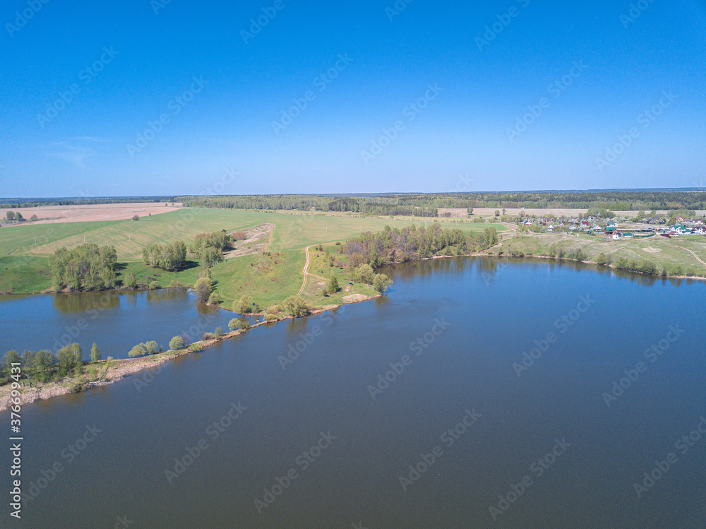 Aerial perspective view blue lake with shore covered by green grass and trees. Drone air view of the beautiful rural landscape with village on background under clear blue sky. Summer sunny day. Aerial perspective view blue lake with shore covered by green grass and trees. Drone air view of the beautiful rural landscape with village on background under clear blue sky. Summer sunny day.