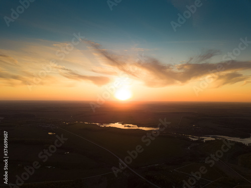 Aerial view of the sunrise over dark rural landscape with lake and river. Rising sun at blue cloudy sky over horizon and dark surface with reflecting water.